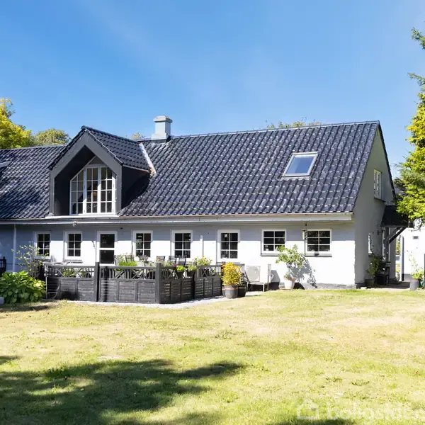 Et hvidt hus med sort tag står i en solrig have med grønt græs og omgivet af træer. Huset har flere vinduer og en lille terrasse med blomsterkasser.