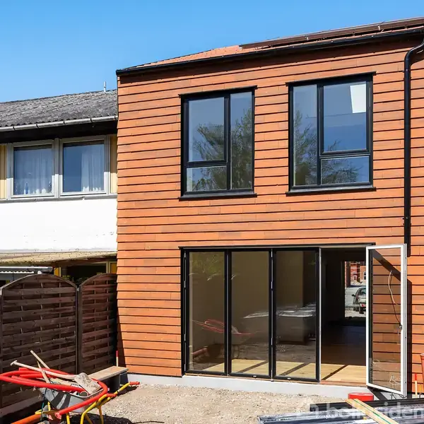 Hus med træbeklædning står under renovering i et boligområde. Byggeværktøjer og materialer ligger på jorden foran, mens nabohuse kan ses i baggrunden.