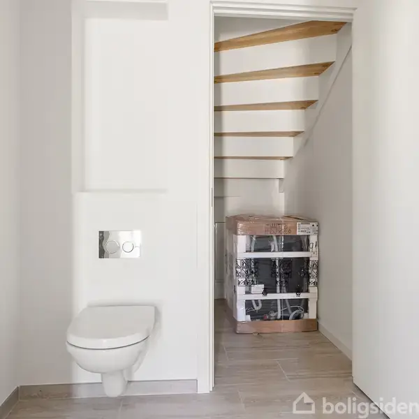 Et lille rum med et væghængt toilet til venstre, uden for brug. Bagerst ses en trappe med lyse trætrin og hvide vægge omkring.