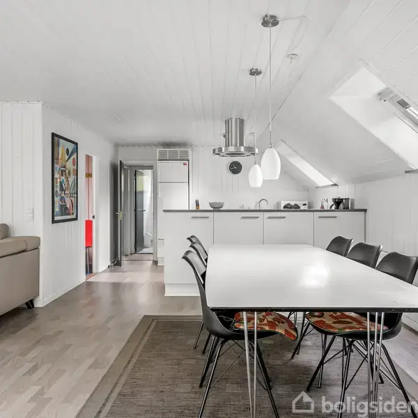 En moderne spisestue med et langt, hvidt bord og sorte stole. Køkkenet er i baggrunden. Rummet er lyst og rummeligt med et par skråvinduer og kunst på væggene.
