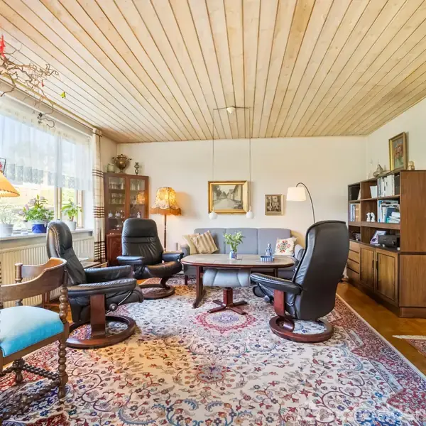 Stue med to sorte lænestole omkring et rundt sofabord, dekoreret med blomster, placeret på et orientalsk tæppe. Rummet har træloft, bogreoler og store vinduer med gardiner.