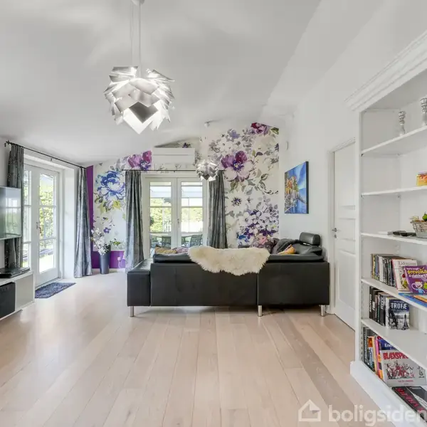 Stue med sort sofa placeret mod en væg med blomstret tapet, belysning fra moderne lampe. TV på møbel i venstre side. Reol med bøger og dekorationer.