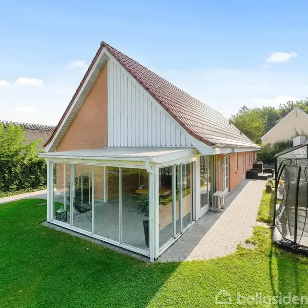 Et moderne hus med rødbrun murstensfacade og rødt tag har en stor udestue med glasvægge. I haven står en trampolin på den grønne græsplæne.