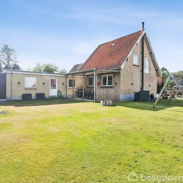 En rødtagget murstenshus står i en grøn have med gyngestativ. Sollys falder på huset, og der er træer i baggrunden.