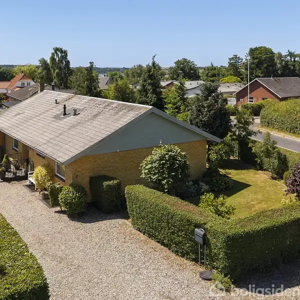 En gul murstensvilla med grå tag, omgivet af velplejet have og hække. Står ved en grusvej på en rolig villavej. Kurvedrev og velholdt forhave med græs og buske.