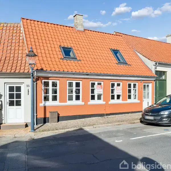 Et rækkehus med orange murstensfacade og rødt tegltag har flere vinduer. En sort bil er parkeret foran. Til venstre ses et hvidt hus med nummer 15 ved døren.