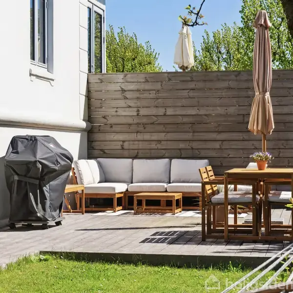 Terrassemøbler står stille på en flisebelagt terrasse, omgivet af en trævæg. En gasgrill er dækket til venstre, og en hængekøje hænger tæt på et træ i en grøn have.