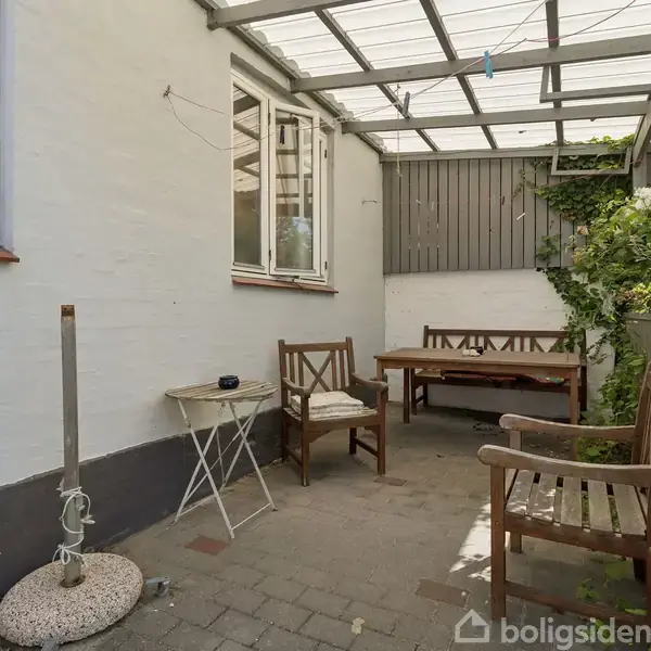 Siddeplads under tag lavet af gennemsigtigt plast, bestående af træbænk og stole ved et bord, placeret på flisebelagt terrasse ved hvidt hus, omgivet af grønne planter.