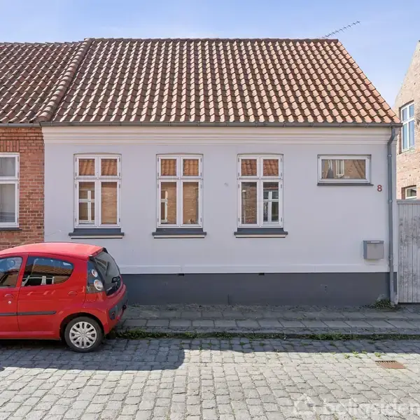 En rød bil er parkeret foran et hvidt hus med teglstenstag. Huset har fem vinduer og en nummer 8 ved døren, placeret på en brostensbelagt gade.