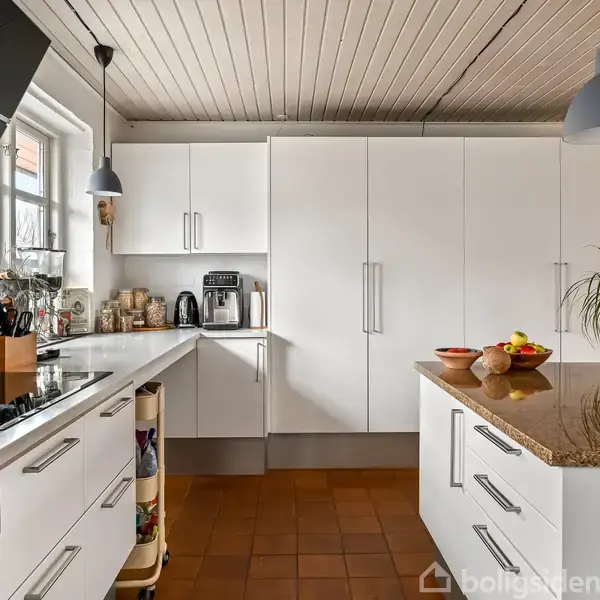 Et moderne køkken med hvide skabe og skuffer. Køkkenudstyr, en kaffemaskine og en plante står på bordpladen. En frugtskål ses på øen. Vinduet lader lys ind.