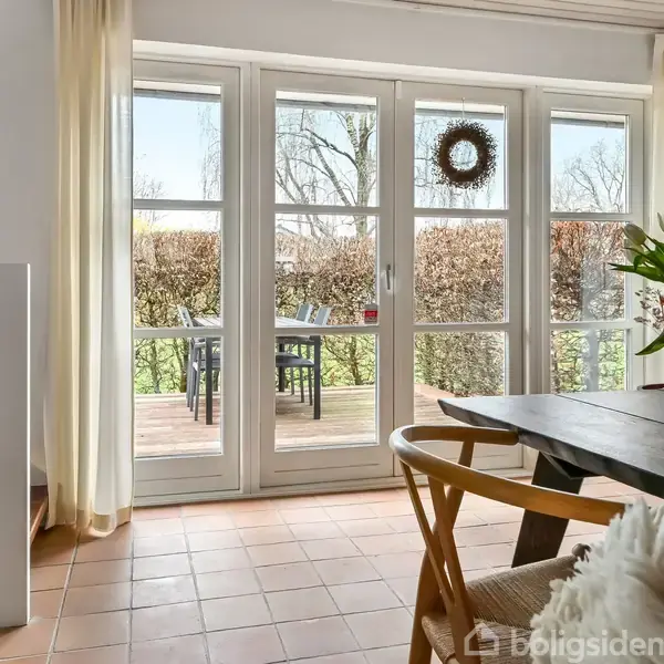 En stue med et trægulv og et stort vinduesparti med udsigt til en terrasse. Et bord med blomster står ved siden af stole. Sollys strømmer ind.