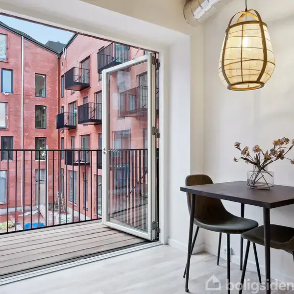 En balkon ses gennem åbne døre ved et lille spisebord med to stole. En hængelampe lyser rummet op. Udenfor er en rød murstensbygning med flere altaner.