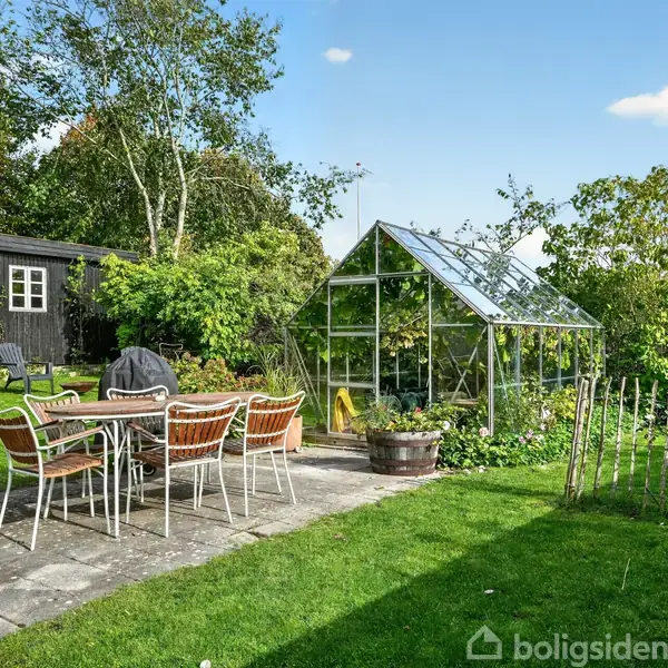 Et havebord med stole står på en flisebelagt terrasse. Ved siden af er et drivhus omgivet af grønne planter og træer. I baggrunden ses et sort skur.