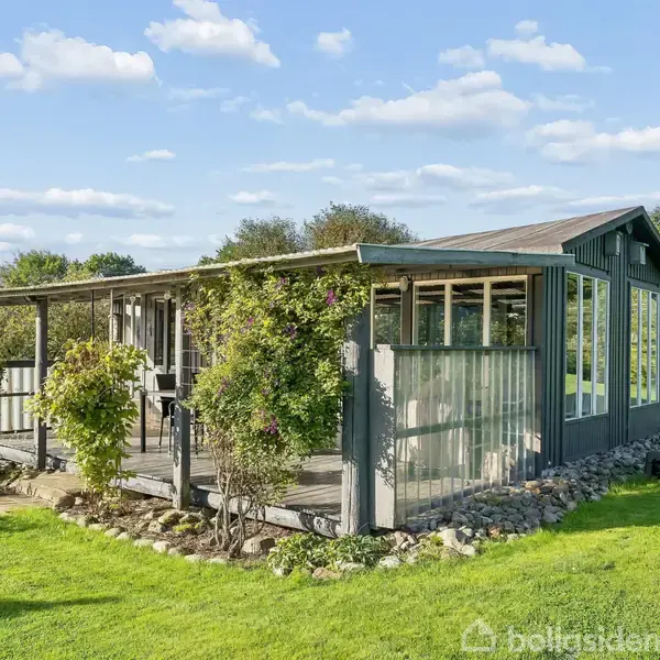 Et lille, gråt sommerhus med store vinduer står omgivet af en frodig have med grønne buske og græsplæne. Sollys oplyser scenen, og himlen er blå med skyer.