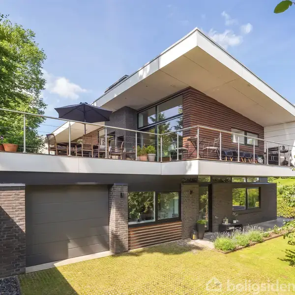 Moderne hus med fladt tag og store vinduespartier. Omkringliggende terrasse med møbler og parasol. Græsplæne og træer omkranser huset. Solskin og blå himmel i baggrunden.