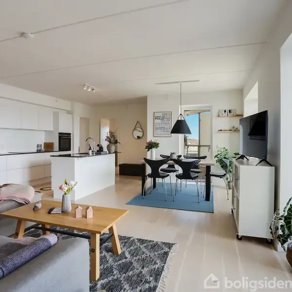 Stue med sofa og sofabord foran et åbent køkken. Spisebord med fire stole står på et blåt tæppe. Store vinduer giver udsigt over omgivelserne. Planter dekorerer rummet.