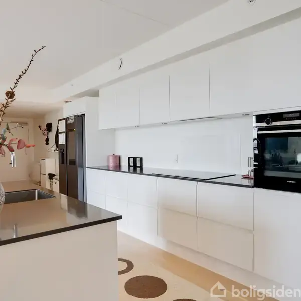 Et moderne køkken med hvide skabe, indbygget ovn og kogeplade. På køkkenøen står en vase med tørrede blomster. Lyse vægge og minimalistisk design præger omgivelserne.