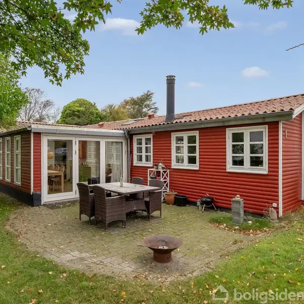 Et rødt træsommerhus med hvide vinduer står omgivet af grønne træer og græsplæne. En havemøbelgruppe står på en fliseterrasse foran huset under en blå, skyfri himmel.