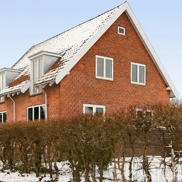 Rødt murstenshus med snedækket tag står stille i et snedækket, villakvarter. Husets facade har flere vinduer og omgives af en hæk og en træbeklædt have.