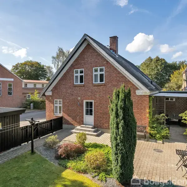 En murstensvilla med skråt tag står i en have med grønne buske og træer. Terrasse med bord og stole ses foran huset under en blå, skyet himmel.