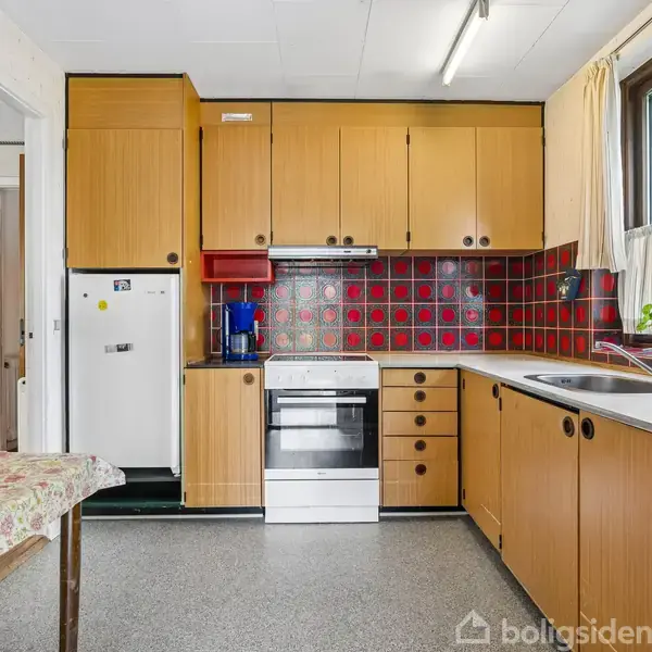 Et køkken med brune træskabe og en ovn i midten. Rødt-og-sort flisebelagt backsplash bag ovnen. Vindue på højre side med udsigt til grønne omgivelser. Bordplade med vask.