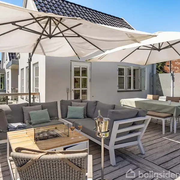 Terrassemøbler står på en træterrasse med grå sofaer, et bord og store hvide parasoller. I baggrunden ses en hvid bygning med vinduer og en grill ved siden af møblerne.