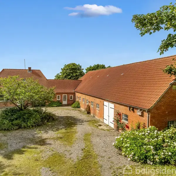 En rød murstensgård med rødt tegltag omgivet af træer og en grusbelagt gårdsplads. Bygningen har flere hvide døre og vinduer, med grønne buske og træer i baggrunden.