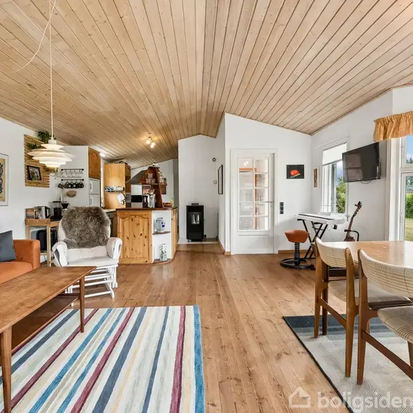 Stue med en træbeklædt loft og gulv, indeholdende en orange sofa, lænestol, sofabord, og spisebord. Store vinduer viser udsigt til have; køkken og komfur i baggrunden.
