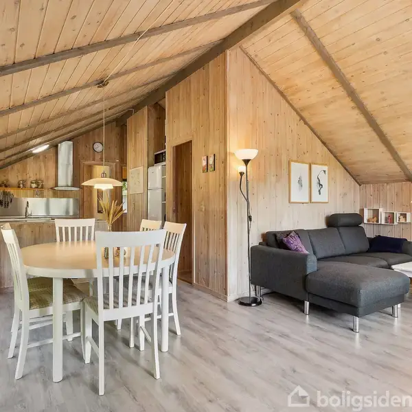 Træbeklædt stue med spisebord og stole. Sofa og sofabord i opholdssektionen. Køkken i baggrunden med rustfrit stål køkkenudstyr. Store vinduer giver udsigt til grønt landskab.
