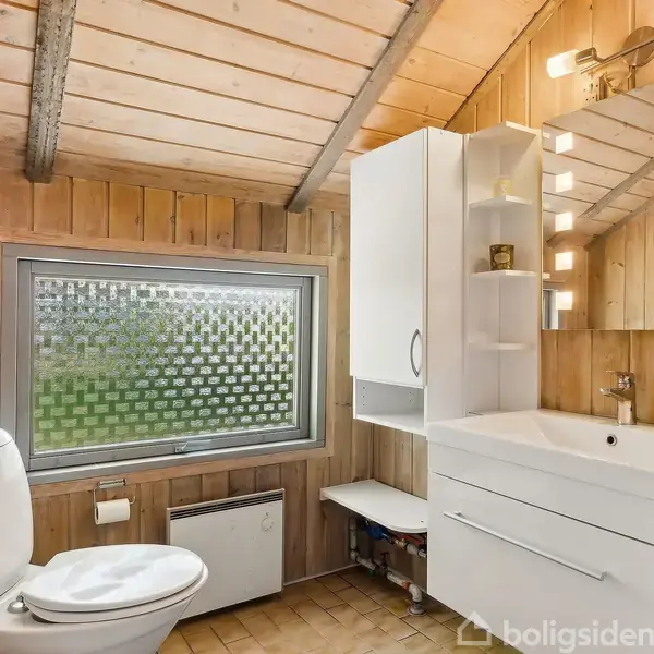 Toilet står stille i træbeklædt badeværelse med skrå loft. Vaskeområde har hvidt skab og spejl med lys. Matteret vindue giver privatliv og udsigt.