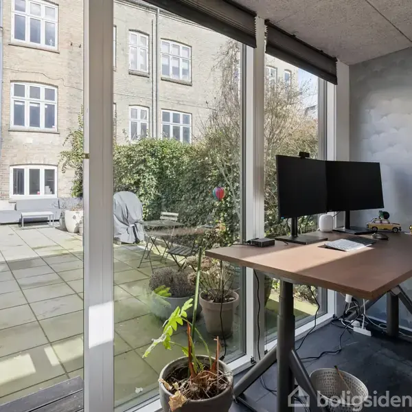 Kontorbord med skærm og planter står tæt ved store vinduer. Udenfor er en flisebelagt gårdhave med havemøbler og planter, omkranset af murstensbygninger med vinduer.