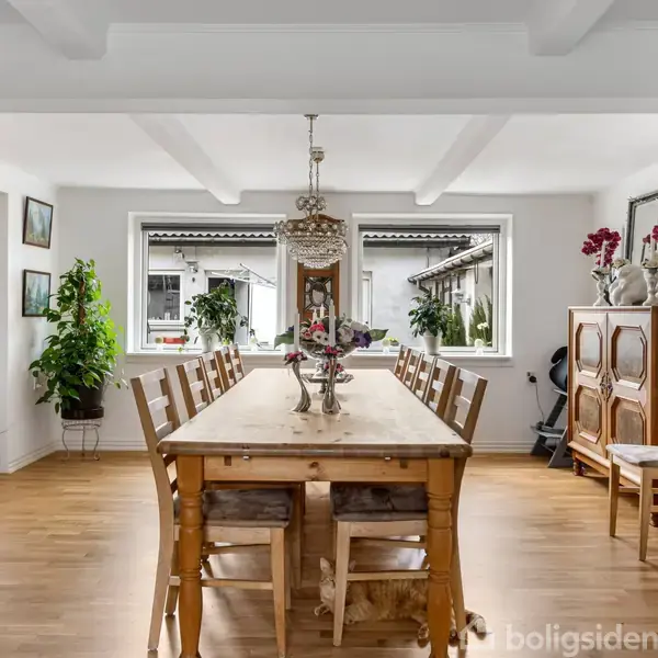 Et træspisebord med stole står midt i en lys spisestue. Rummet har trægulve, hvide vægge og vinduer med udsigt til en have. Dekorationer omfatter planter og lamper.