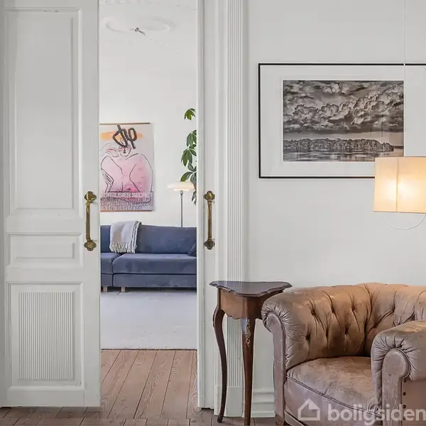 En brun lænestol står ved siden af et lille træbord i et værelse med hvidmalede vægge og trægulv. Gennem en åben dør ses en blå sofa og kunstværker.