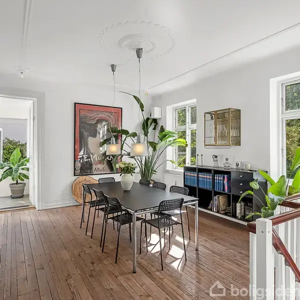 Spisestue med sort bord og seks stole i en lys, rummelig lejlighed. Store vinduer giver naturligt lys. Grønne planter og kunst pryder rummet, der har trægulv og hvide vægge.
