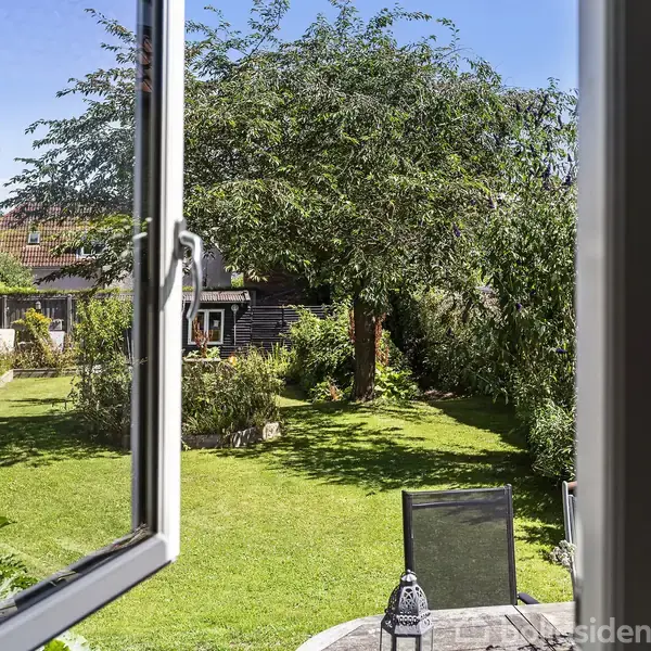 Åbent vindue vender mod en velplejet have, hvor et stort træ skygger græsplænen. Havemøbler og en pool ses i baggrunden under en klar blå himmel.