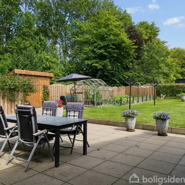 Terrassemøbler står på en flisebelagt terrasse i en have med grøn plæne og træhegn. Et drivhus og nogle blomsterkasser ses i baggrunden omgivet af frodig vegetation.