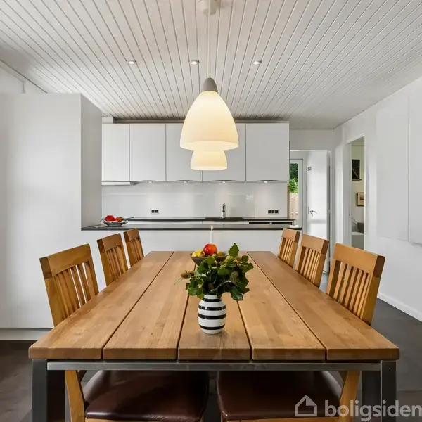 Træbord med seks stole står centralt, dekoreret med en vase med blomster. Baggrunden har et åbent køkkenområde med lyse elementer og en hængende lampe.