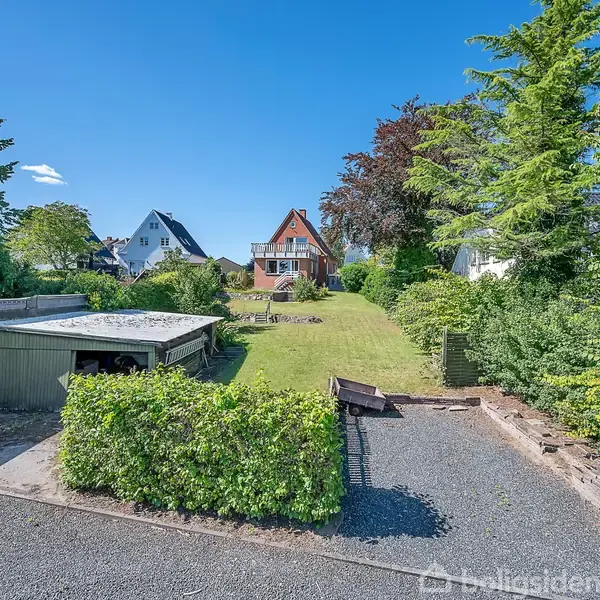 Et hus med en karakteristisk trekantet gavl står i midten. Haven har en græsplæne omgivet af træer og grønne buske. Foran er en grusbelagt indkørsel med en garage.