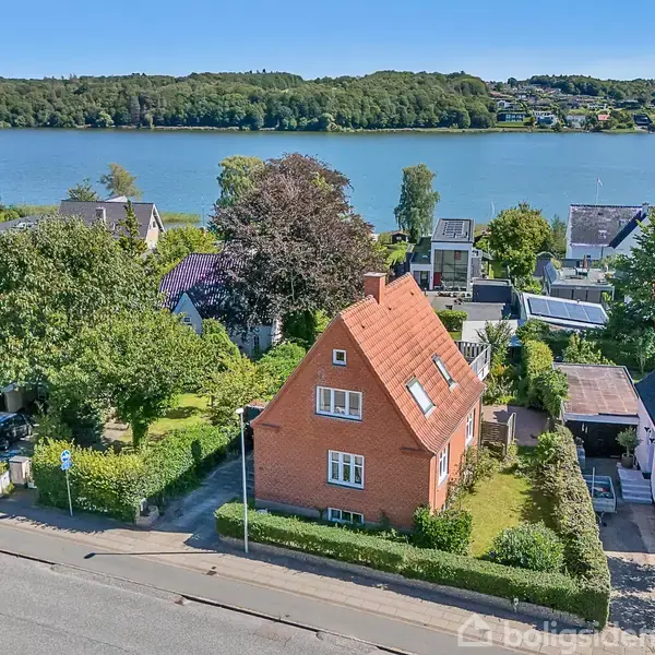 Et rødt murstenshus med skråtag ligger i en grøn have omgivet af andre huse. I baggrunden ses en bred flod og skovklædte bakker under en klar blå himmel.