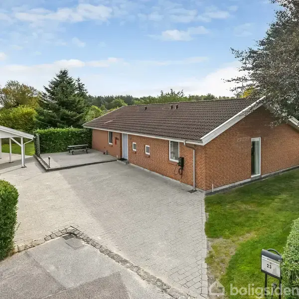 Et udendørs område med en grå flisebelagt terrasse foran. En hvid og grøn rutsjebane står til højre. Baggrunden består af græsplæne og store, grønne træer.