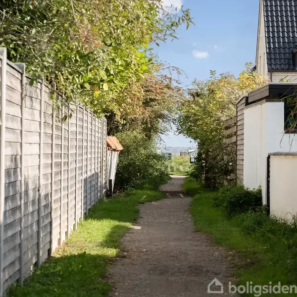 En smal sti løber mellem en hæk og et træhegn, flankeret af et hus til højre. Skyfri himmel og frodig grøn vegetation skaber en fredfyldt landsby-stemning.