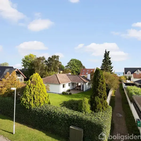 Hvide parcelhuse med have og omkranset af hække. En smal vej løber langs siden, med flere huse og træer i baggrunden under en blå himmel med spredte skyer.