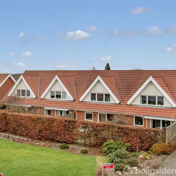 Rækkehus med rødt tegltag og hvide gavle står i en velholdt have med hæk og flagstang. Der er klare, blå himmel og græsplæne i forgrunden.