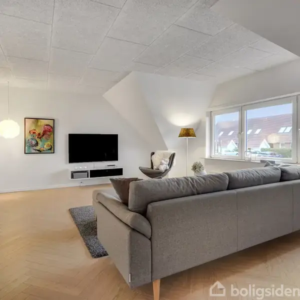 En grå sofa står i en moderne stue med et fladskærms-tv på væggen. Rummet har store vinduer mod et rækkehusområde, en farverig plakat, og designerlamper.