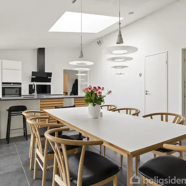 En spisebord med en blomsterbuket står i midten af et moderne køkken og spiseområde. Der er pæne, træstole, et lyst loft med lamper og et køkkenområde med barstole i baggrunden.