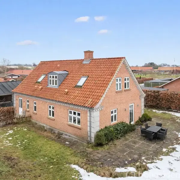 Et rødt murstenshus med rødt tegltag står stille i en snedækket have med flisebelagt terrasse og sorte havemøbler. Andre huse og åbne marker ses i baggrunden.