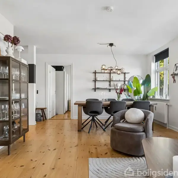 En stue med et spisebord omgivet af stole. En glasvitrine står til venstre. Store planter findes ved vinduet, og belysningen hænger over bordet. Trægulv og hvide vægge kompletterer rummet.
