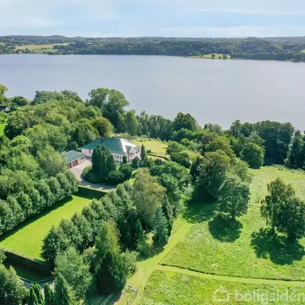 Stort hus omgivet af frodige træer ligger ved en sø. Omkringliggende natur inkluderer grønne marker og skovområder, der strækker sig hen til søens bred.