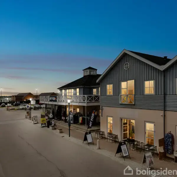 En række bygninger med oplyste vinduer står langs en bred gade. Foran dem er borde og skilte. Biler er parkeret langs vejen, og himlen er mørkeblå ved skumringstid.