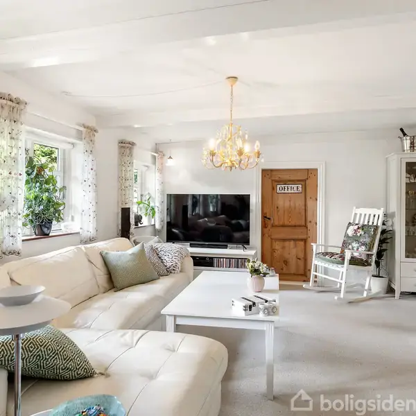 En hvid sofa står i en lys stue ved siden af store vinduer. En stor TV-skærm er placeret på et møbel, og der hænger en lysekrone over et sofabord. Dør med skiltet "Office".